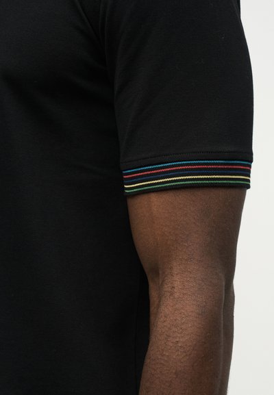 Χέρι ατόμου που φοράει μαύρο κοντομάνικο πουκάμισο με πολύχρωμη ριγωτή μανσέτα στο μανίκι, πάνω σε απλό φόντο.