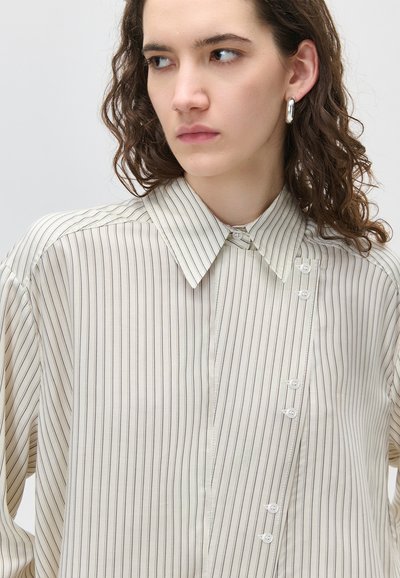 Fiatal nő göndör barna hajjal, bézs csíkos gombos ingben, ezüst karika fülbevalóval, egyszínű háttér előtt.