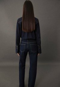 Dunkelblaue Jeanshemd und Jeans, beide im klassischen Schnitt. Das Hemd hat Knopfmanschetten, während die Jeans dekorative Nähte an den Taschen aufweisen.