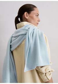 Lichtblauwe gebreide sjaal gedrapeerd over een crème corduroy overhemd met geribbelde textuur. De sjaal heeft een gladde afwerking en zachte stof.