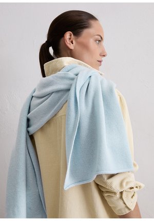 Écharpe tricotée bleu clair drapée sur une chemise en velours côtelé crème avec une texture côtelée. L'écharpe présente une finition lisse et un tissu doux.