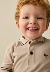 Polo beige avec un tissu texturé, boutons noirs et accents bleus sur le col. Présente une petite broderie d'un animal brun sur la poitrine.
