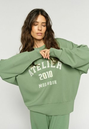 Frau mit langem braunem Haar, die einen locker sitzenden grünen Pullover mit der Aufschrift "ATELIER 2010 MOS MOSH" und passende Hosen trägt und mit verschränkten Händen posiert.