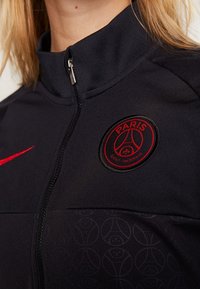 Gros plan sur une veste noire du Paris Saint-Germain avec un logo rouge brodé et un swoosh rouge de Nike sur la poitrine, col partiellement zippé.