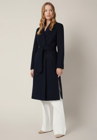 Granatowy długi płaszcz o fasonie kopertowym, z paskiem w talii i dwoma bocznymi kieszeniami. Stylizowany na białych szerokich spodniach, charakteryzujących się gładką fakturą.
