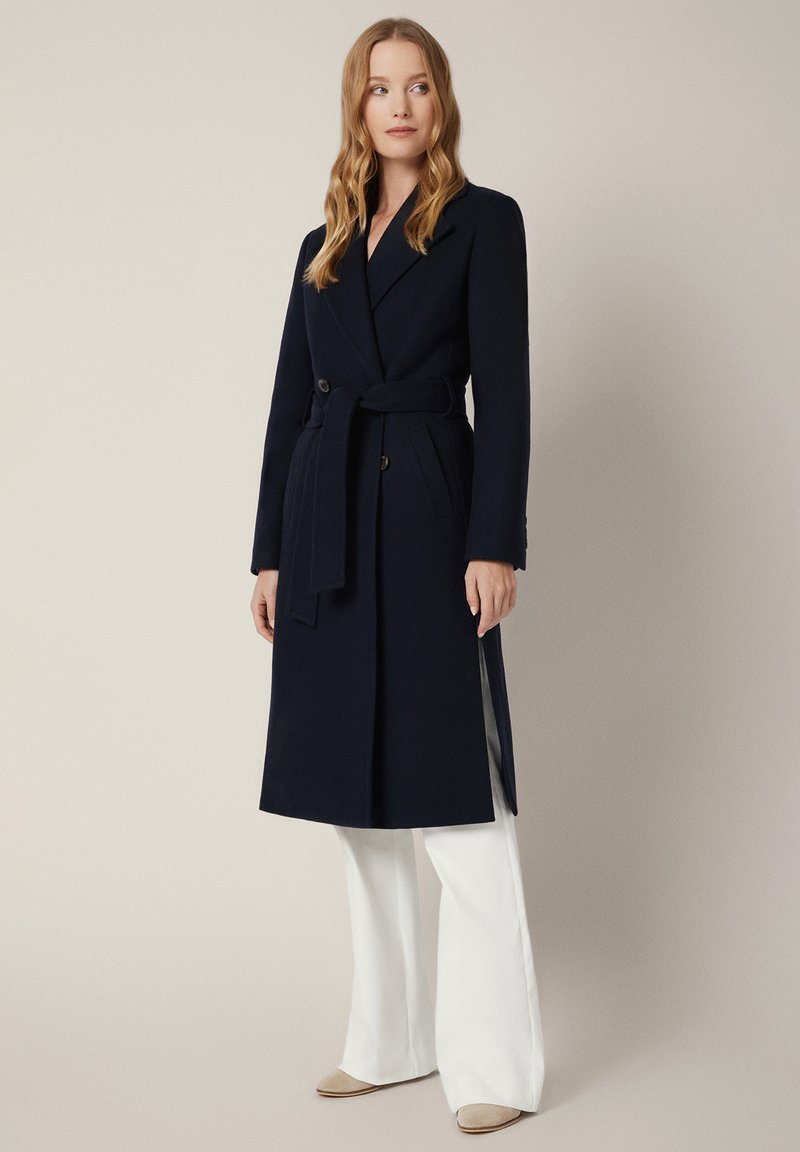 Granatowy długi płaszcz o fasonie kopertowym, z paskiem w talii i dwoma bocznymi kieszeniami. Stylizowany na białych szerokich spodniach, charakteryzujących się gładką fakturą.