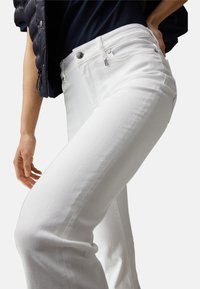 Le jean en denim blanc présente une coupe ajustée avec des coutures discrètes, une poche avant et un bouton argenté à la ceinture.