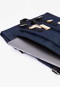Granatowa torba na laptopa o teksturowanej tkaninie; z elementami skórzanymi, regulowanymi paskami i widocznym częściowo w kieszeni zewnętrznej laptopem w kolorze srebrnym.