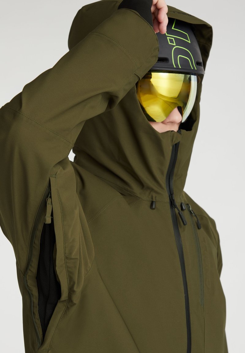 Grüne technische Jacke mit hohem Kragen, Reißverschlussbelüftungen und schwarzen Akzenten. Gelb getönte Skibrille und ein schwarzer Helm werden getragen.