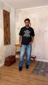 Άνδρας με dreadlocks που φορά μαύρο t-shirt με άσπρο κείμενο, στέκεται σε ένα δωμάτιο με χαλί με σχέδια, ξύλινη καρέκλα και καφέ δερμάτινη τσάντα.