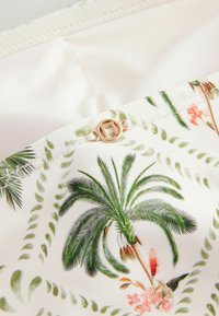 Tissu à motif floral présentant des feuilles de palmier vertes et des fleurs roses sur un fond crème. Détail de bouton en or rose sur une couture.