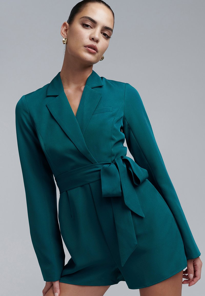 Sini-vihreä pitkähiha kietaisumekko, jossa on vyötäröllä vyö, istuva leikkaus, terävät kaulukset ja sileä pinta. Sopii virallisiin tai puoli-virallisiin tilaisuuksiin.