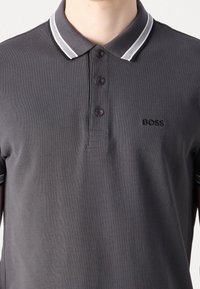 Homme portant un polo gris foncé avec un col à rayures blanches et le logo noir "BOSS" sur la poitrine gauche.