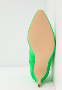 Chaussure à talon vert posée sur la semelle contre un coin blanc, montrant une semelle beige lisse et une fine pointe de talon.