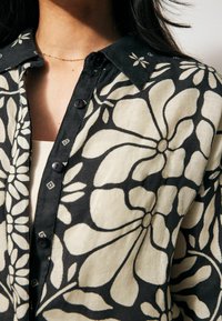 Koszula z guzikami z kwiatowym wzorem w czerni i kremie, wykonana z fakturowanej tkaniny, z kołnierzykiem i czarnymi guzikami.