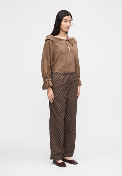 Kvinne iført brun semsket bluse med rysjekanter på ermene og mønstrete vide bukser, stående med hendene avslappet langs siden.