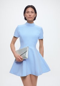 Γυναίκα με κοντομάνικο, ανοιχτό μπλε φόρεμα που κρατάει ασημένιο τσαντάκι αντίθετα σε λευκό, απλό φόντο.