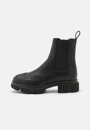 Botte Chelsea en cuir noir avec perforations brogue, panneaux latéraux élastiques, languette de tirage et semelle épaisse à crampons, vue de profil.