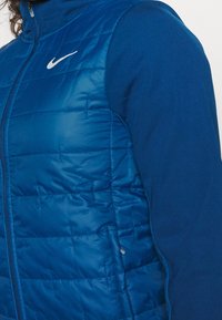 Veste sans manches matelassée bleue avec un design matelassé, des manches bleues contrastantes et un logo Nike blanc sur la poitrine. Fermeture éclair et tissu léger.
