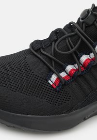 Zwarte sportieve schoen met een gestructureerde mesh bovenkant, elastisch vetersysteem en kleurrijke streeppatroon in rood, wit en blauw.
