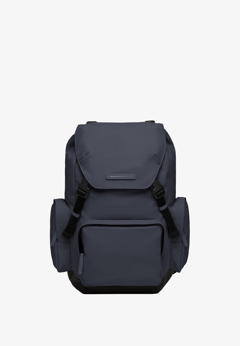 Sac à dos gris en tissu durable, équipé d'une fermeture à rabat, de bretelles réglables, d'une poche frontale et de poches latérales pour un rangement supplémentaire.