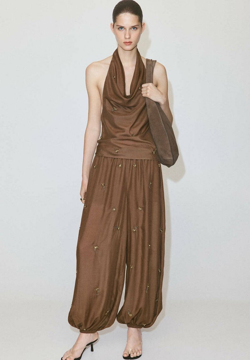 Femme portant un top marron sans manches style halter et un pantalon ample assorti avec de petits détails floraux, tenant un grand sac à épaule marron.