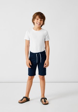 Weißes T-Shirt mit kurzen Ärmeln, marineblaue Shorts mit Kordelzug und schwarze Flip-Flops. Einfaches Design mit bequemer Passform und weicher Textur.
