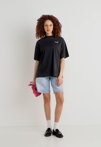 Γυναίκα με σγουρά μαλλιά στέκεται φορώντας ένα μαύρο, φαρδύ t-shirt, ανοιχτόχρωμα denim σορτς, λευκές κάλτσες, μαύρα loafers και κρατώντας μια μικρή κόκκινη τσάντα.