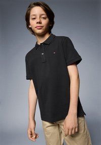 Polo shirt nera con colletto, maniche corte e logo sul petto. Abbinata a pantaloni beige. Texture liscia e design classico.