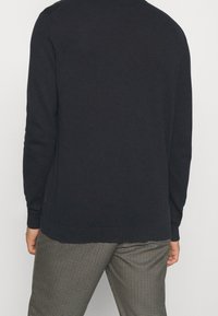Maglione girocollo di un blu scuro con maniche lunghe, polsini e orlo a costine. Texture liscia, branding minimale sul lato inferiore. Abbinato a pantaloni a quadri.