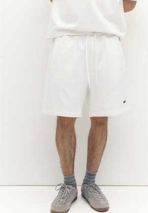 Muž v bielych teplákových šortkách so sťahovacou šnúrkou, sivé pruhované ponožky a sivé tenisky, stojí pred jednoduchým bielym pozadím.