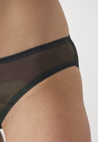 Mørkegrønn mesh undertøy med scalloped kant og kontrasterende svart trim. Materialet er semitransparent med en glatt tekstur.