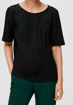 Kobieta w czarnej bluzce z krótkim rękawem i plisami przy dekolcie oraz w ciemnozielonych spodniach, stojąca z jedną ręką w kieszeni.