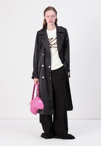 Cappotto lungo nero con bottoni argentati, indossato sopra una maglietta bianca stampata e pantaloni neri a gamba larga. Tiene una borsa lucida rosa.