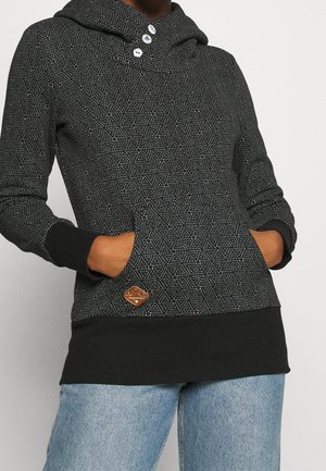 Persoon die een zwarte hoodie met patroon draagt met voorzakken, drie knopen bij de hals en lichtblauwe spijkerbroek zichtbaar vanaf halverwege het dijbeen naar beneden.
