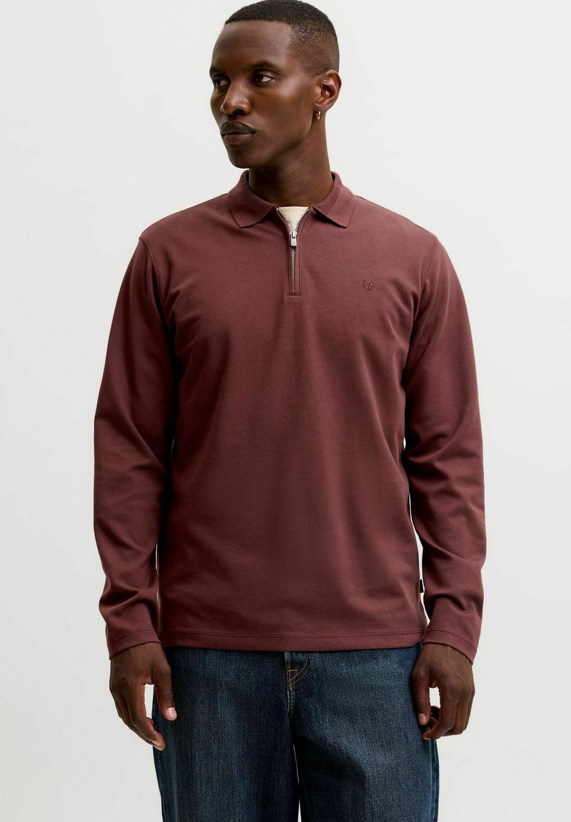 Homme portant un polo bordeaux à manches longues avec un col zippé et un jean bleu, debout devant un fond clair uni.