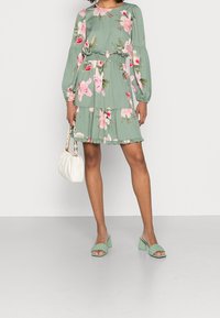 Robe verte fleurie à manches longues avec volants en étages. Associée à des sandales à talons blocs vert clair et un sac à main blanc.