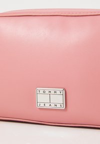 Rosa syntetisk läderväska med slät yta, med en rektangulär metall-etikett som står "TOMMY JEANS" på sidan.