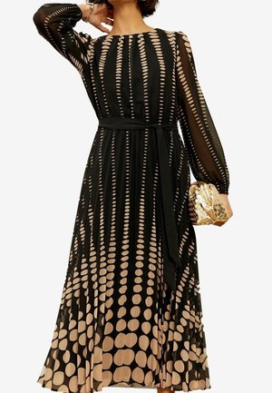 Kvinde iført en sort kjole med lange ærmer med beige cirkelmønstre, et bælte i taljen og holder en skinnende gylden flettet clutch.
