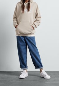 Ljusbeige huvtröja med ficka framtill och vit "adidas SPORTSWEAR"-logotyp, matchad med lösa blå denimjeans och vita sneakers.