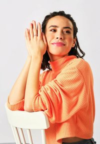 Ribbet turtleneck sweater i lys orange med løse ærmer. Modellen poserer med hænderne tæt på ansigtet og viser en delikat ring på fingeren.