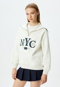 Sudadera blanca con media cremallera, cuello y puños de canalé. Presenta texto impreso en azul "NYC NEW YORK CITY 1998 USA 2001". Tejido texturizado.