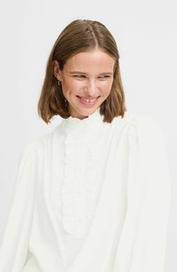 Hvid bluse med en høj flæsekrave, knaplukning og lange, voluminøse ærmer. Stoffet ser blødt og letvægtigt ud.