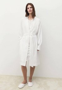 Halat de baie alb cu mâneci lungi, talie cu curea și buzunare laterale; suprapus peste o rochie albă. Material moale, design simplu, detalii minime.