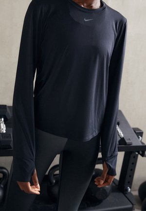 Person wearing black long-sleeve Nike workout top and leggings standing in front of gym equipment, including kettlebells and dumbbells.