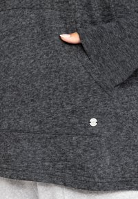 Gray textured hoodie fabric with a front pocket, featuring a metallic accent. The design includes thumb openings in the cuffs.