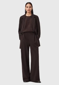 Donna con capelli scuri e lisci indossa un blazer marrone scuro, un maglione soffice e pantaloni a gamba ampia, in piedi con le mani nelle tasche su uno sfondo neutro.
