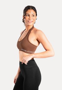 Brązowy sportowy biustonosz prążkowany z wiązaniem na szyi i białym logo "SMILODO". W połączeniu z czarnymi legginsami z wysokim stanem. Gładka faktura.