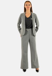 Blazer grigio con revers e chiusura a bottone, abbinato a pantaloni larghi grigi e una maglietta a righe nera, indossato con tacchi neri lucidi.