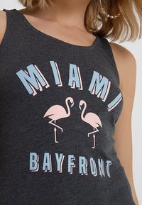 Szary top bez rękawów z napisem "MIAMI BAYFRONT" w jasnoniebieskim i różowym kolorze, z dwoma stylizowanymi różowymi flamingami umieszczonymi centralnie poniżej.